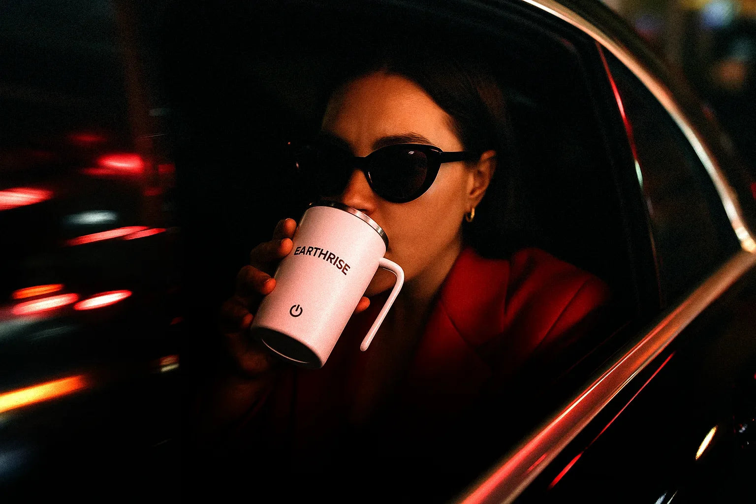 A woman drinking out of an Earthrise mug, sitting in the backseat of a car