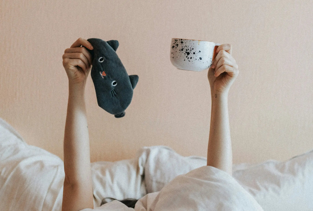 Two arms rising from a bed one holding a mug the other holding a cat shaped eye mask with beige background